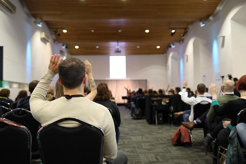 A crowd watching a presentation, man in front of camera raising hand to ask question. 